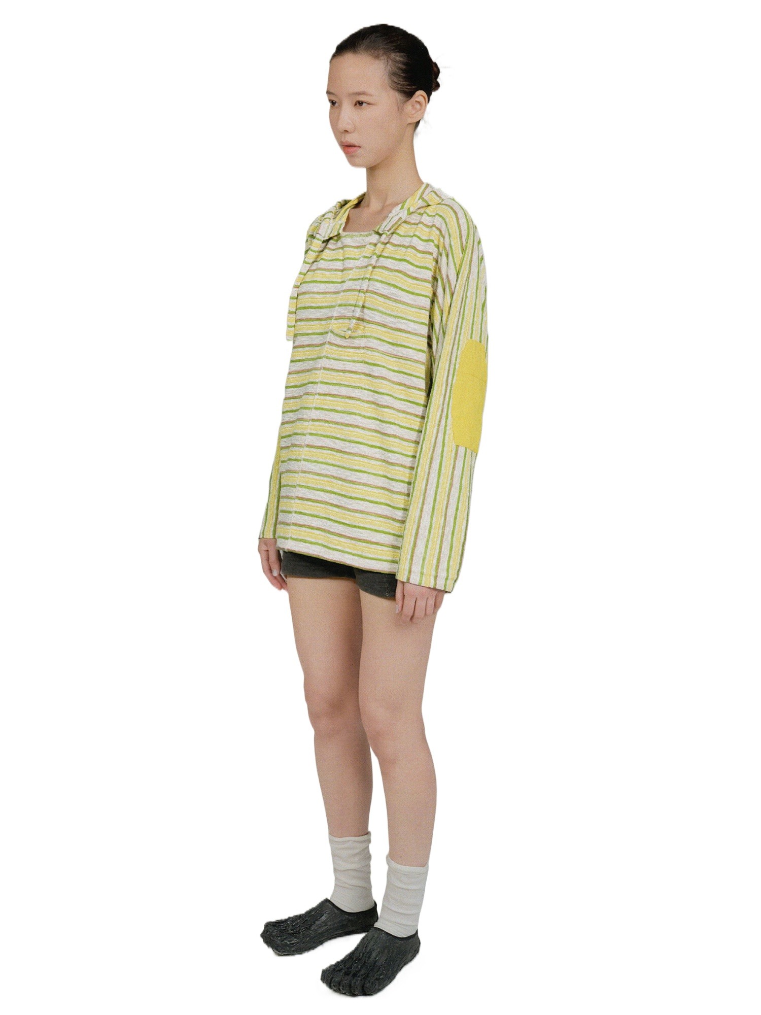 一个人面向东方,身穿 EastLyst 出品的巴哈风格黄绿条纹针织套头衫、黑色短裤、灰色袜子和黑色鞋子。头发扎在脑后,站在纯白色背景前。