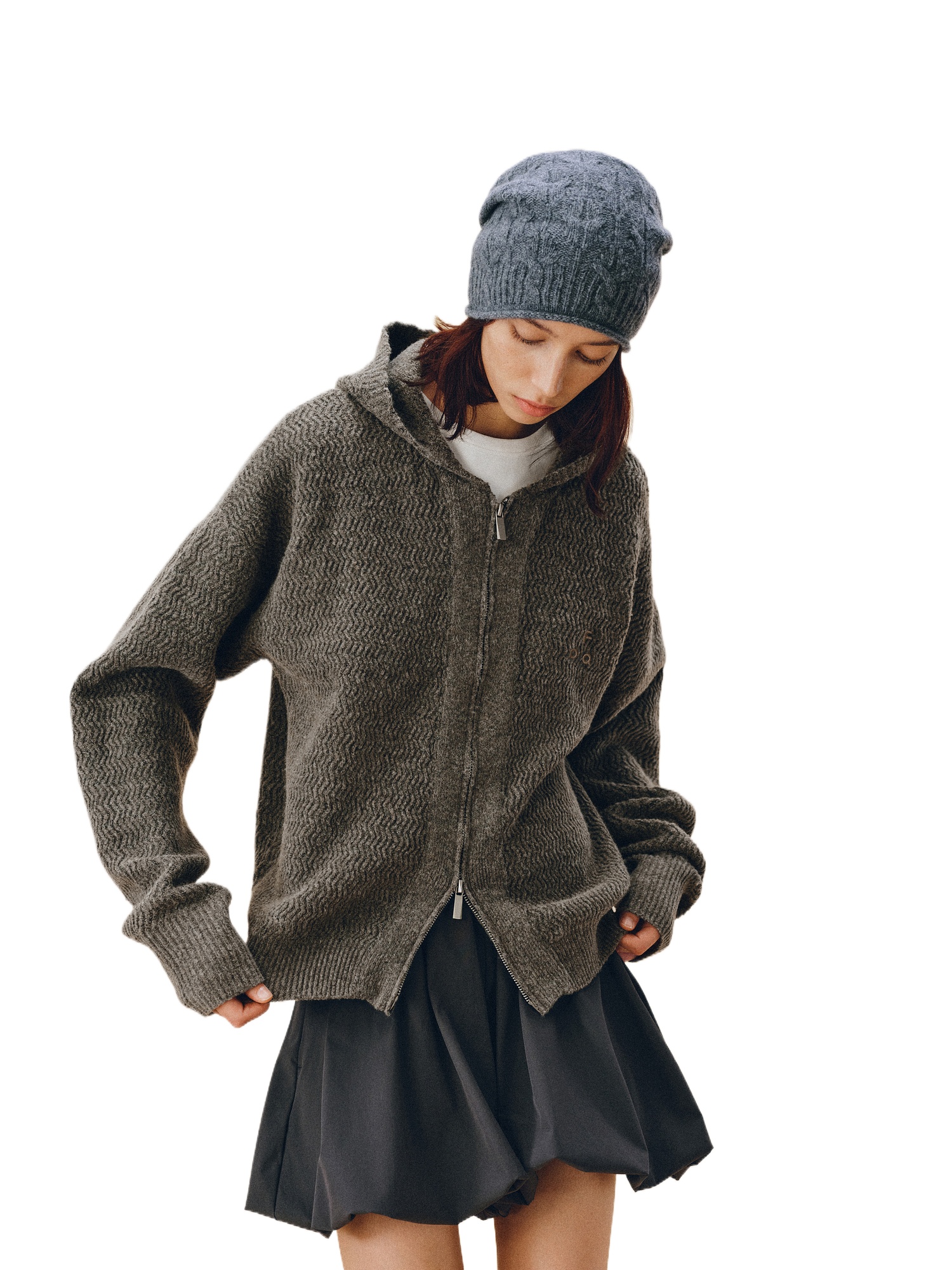 Wearing an EastLyst Color-Block Hooded Cardigan, a matching knit beanie, and a black pleated skirt, the person stands with hands in pockets and looks down against a white background.
